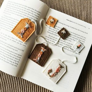 May include: A collection of handmade felt bookmarks with floral embroidery. The bookmarks are in various shapes and colors, including brown, tan, and white, with delicate floral designs in pink, orange, and green. They are arranged on an open book.
