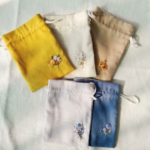 Puede incluir: Cinco pequeñas bolsas de tela con cordón de ajuste y diseños florales bordados. Las bolsas son de varios colores, incluyendo amarillo, beige, blanco, rosa y azul.