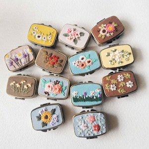 Peut inclure: Une collection de boîtes métalliques rectangulaires à charnières, chacune ornée de broderies florales complexes. Les boîtes présentent divers coloris, dont le jaune, le rose et le bleu, avec des motifs de fleurs cousues. Les boîtes sont disposées sur une surface blanche.