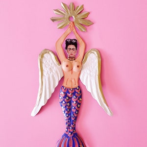 Puede incluir: Un adorno de metal colorido que representa una sirena con alas. La sirena tiene un torso humano con un retrato pintado de Frida Kahlo y una cola de pez azul y roja. El adorno está coronado con una estrella dorada.