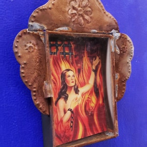 May include: A framed image of a woman in a fiery setting, with a rusted metal frame featuring a decorative flower design.