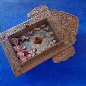 May include: A small, ornate metal frame with a heart-shaped image inside. The frame is decorated with intricate designs and has a distressed, aged look. The image inside the frame depicts a heart with a crown of thorns and angels surrounding it. There are also small pink flowers around the image.