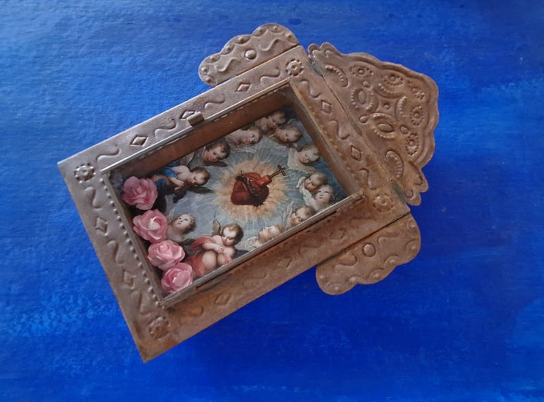 May include: A small, ornate metal frame with a heart-shaped image inside. The frame is decorated with intricate designs and has a distressed, aged look. The image inside the frame depicts a heart with a crown of thorns and angels surrounding it. There are also small pink flowers around the image.