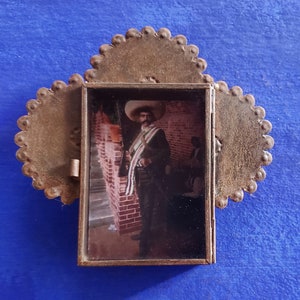 May include: A small, rustic metal frame with a scalloped edge, containing a black and white photograph of a man in a sombrero and a sash. The man is standing in front of a brick wall.