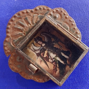 K&ouml;nnte beinhalten: Ein kleiner, verzierter Metallrahmen mit rostiger Oberfl&auml;che, der ein Vintage-Bild eines Engels mit Fl&uuml;geln enth&auml;lt. Der Rahmen hat einen gewellten Rand und ein dekoratives Design.