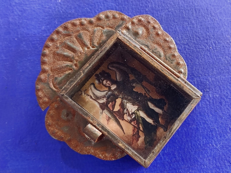 K&ouml;nnte beinhalten: Ein kleiner, verzierter Metallrahmen mit rostiger Oberfl&auml;che, der ein Vintage-Bild eines Engels mit Fl&uuml;geln enth&auml;lt. Der Rahmen hat einen gewellten Rand und ein dekoratives Design.