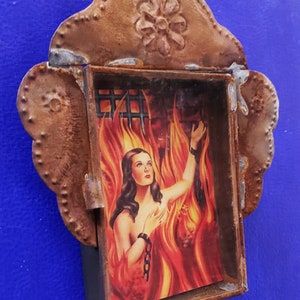 May include: A rusty metal frame with a decorative flower design on top. Inside the frame is a picture of a woman with long dark hair, wearing a chain around her wrist, standing in a fiery inferno.
