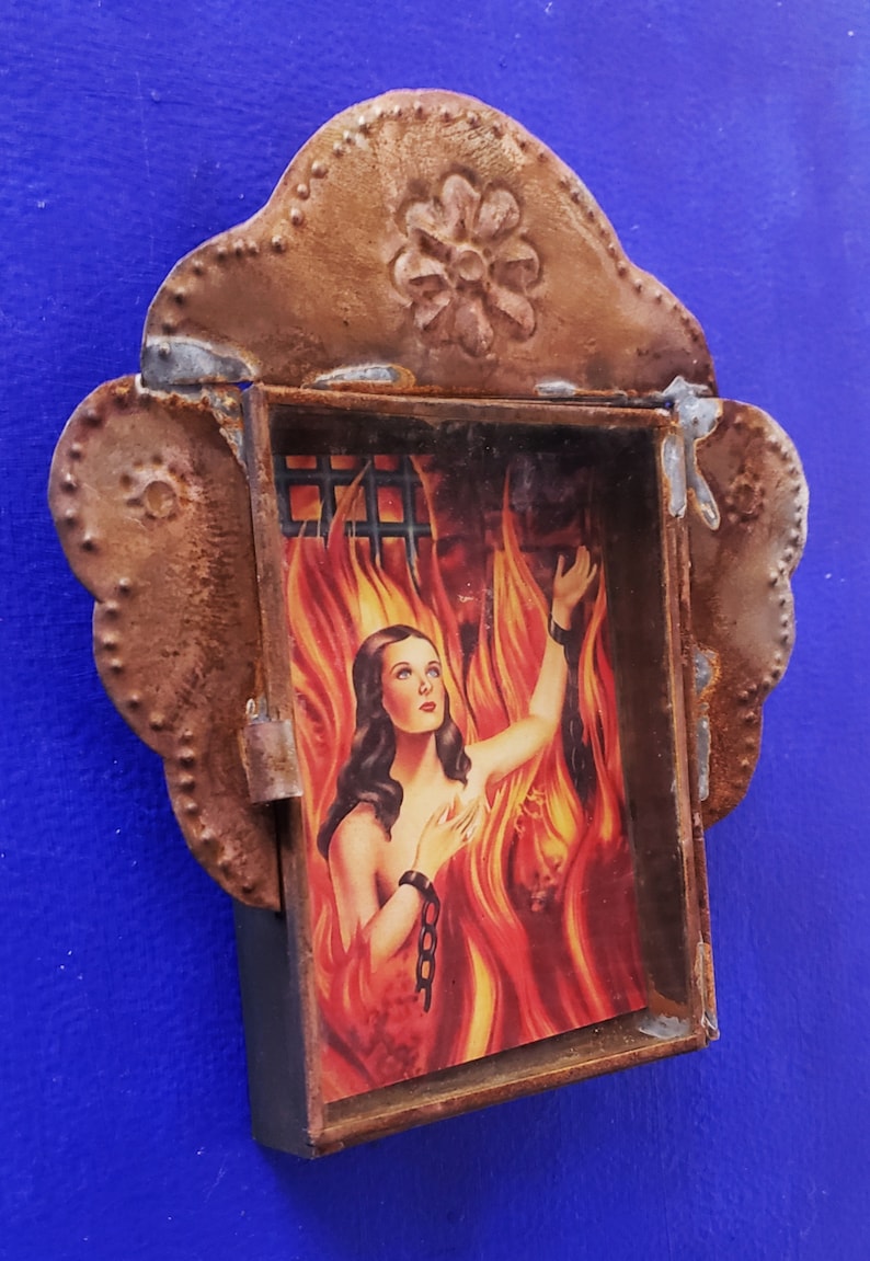 May include: A rusty metal frame with a decorative flower design on top. Inside the frame is a picture of a woman with long dark hair, wearing a chain around her wrist, standing in a fiery inferno.