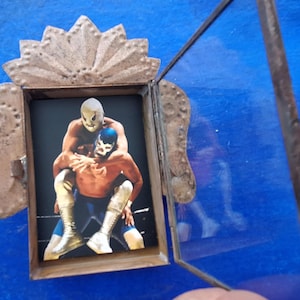 May include: A framed picture of two wrestlers in a wrestling match. The wrestlers are wearing masks and are in a fighting pose. The frame is made of wood and has a glass front.