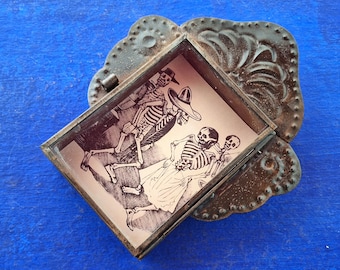 May include: A small, ornate metal box with a hinged lid. The box contains a printed image of three skeletons in a procession, with one wearing a sombrero. The box is decorated with a floral design and has a rusty patina.