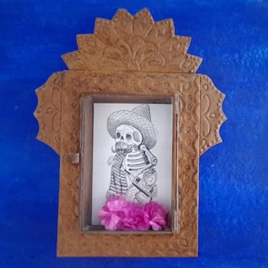 May include: A small, ornate, metal frame with a decorative design. The frame contains a black and white drawing of a skeleton wearing a sombrero and holding a guitar. Pink flowers are placed at the bottom of the frame.