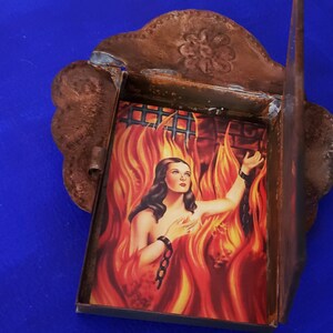 May include: A small, rusty metal frame with a hinged door, containing a vintage image of a woman in flames. The image is set against a backdrop of a brick wall and a barred window.