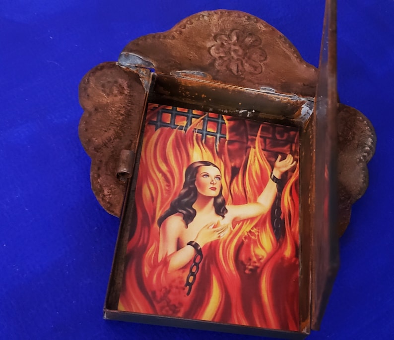 May include: A small, rusty metal frame with a hinged door, containing a vintage image of a woman in flames. The image is set against a backdrop of a brick wall and a barred window.