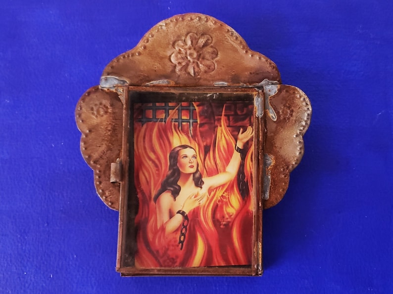 May include: A small, ornate metal frame with a glass window containing a vintage image of a woman in flames. The frame is decorated with a flower design and has a scalloped edge.