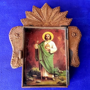 May include: A small, ornate, wooden frame with a hinged door, featuring a religious image of a man in a green robe holding a staff. The frame is decorated with a sunburst design and has a gold-colored finish.