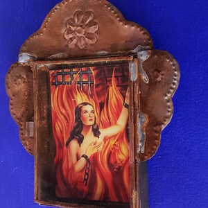 May include: A framed image of a woman in a fiery setting, with a chain around her wrist. The frame is made of metal with a decorative floral design at the top.