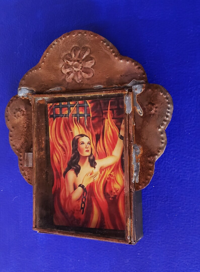 May include: A framed image of a woman in a fiery setting, with a chain around her wrist. The frame is made of metal with a decorative floral design at the top.