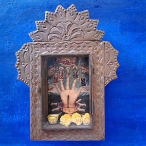 May include: A small, ornate, metal frame with a hand and figures inside. The frame is decorated with a floral design and has a blue background.