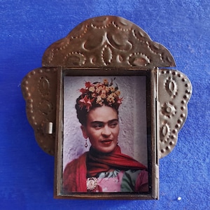 May include: A framed picture of Frida Kahlo with a floral crown, wearing a red scarf and a green and pink dress. The frame is made of metal with a decorative design.