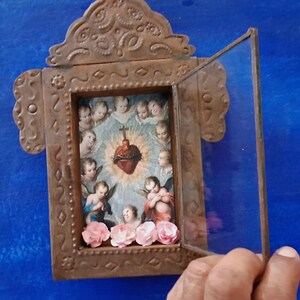 May include: A small, ornate, metal frame with a glass door that opens. Inside the frame is a religious image of a heart with thorns and angels surrounding it. The frame is decorated with flowers.