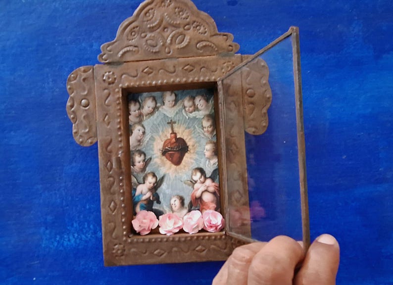 May include: A small, ornate, metal frame with a glass door that opens. Inside the frame is a religious image of a heart with thorns and angels surrounding it. The frame is decorated with flowers.