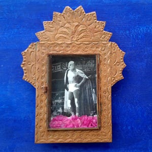 May include: A small, ornate, gold-colored metal frame with a picture of a masked wrestler inside. The frame has a decorative design with a floral pattern at the top. Pink flower petals are placed in front of the picture.