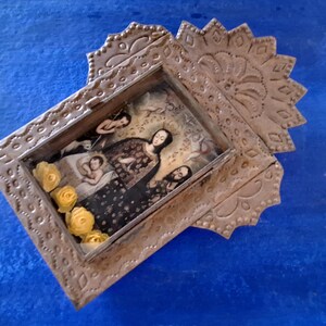 May include: A silver metal frame with an ornate design, featuring a religious image of a woman holding a child. The frame is decorated with yellow flowers.