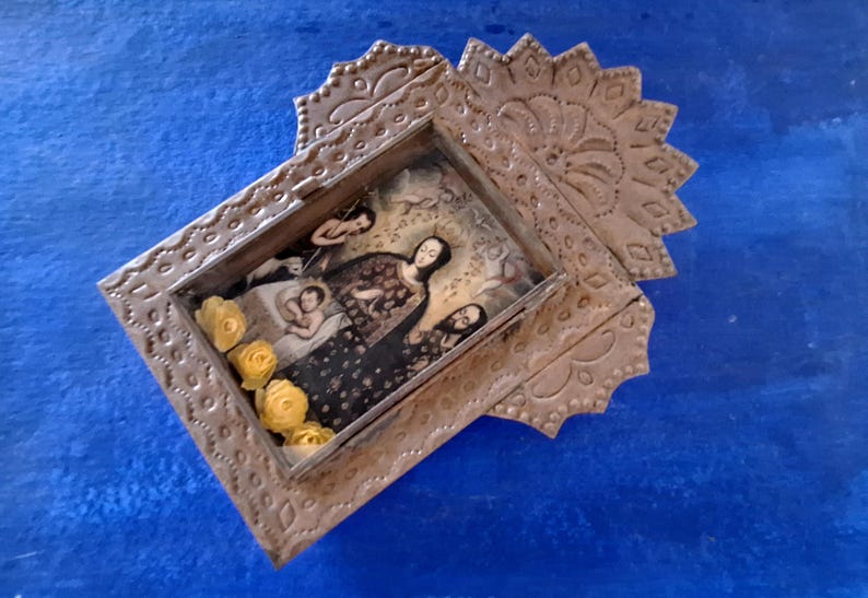 May include: A silver metal frame with an ornate design, featuring a religious image of a woman holding a child. The frame is decorated with yellow flowers.