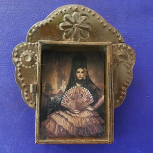 Op de afbeelding: Een foto met een metalen lijst en een decoratief bloemmotief bovenaan. De foto toont een vrouw in een zwarte kanten jurk die een waaier vasthoudt. De achtergrond is een stadsbeeld.