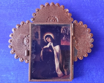 May include: A small, rusty metal frame with a hinged door, containing a printed image of a woman in a white robe and a halo, holding a book. The frame has a scalloped edge and is decorated with small, round rivets.
