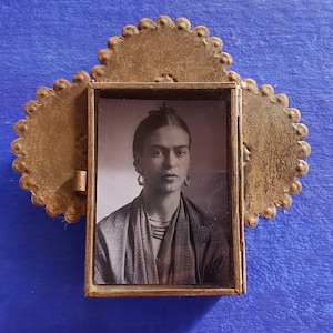 May include: A small, ornate, gold-colored metal frame with a black and white photo of Frida Kahlo inside. The frame has a scalloped edge and is reminiscent of a traditional Mexican retablo.