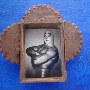May include: A small, rustic, brown metal frame with a scalloped edge, holding a black and white image of a masked wrestler with arms crossed. The frame is set against a blue background.
