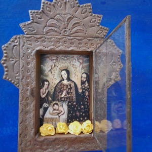 May include: A small, ornate wooden frame with a glass door. The frame is decorated with intricate metalwork and features a religious image of a woman and two children. The image is surrounded by yellow flowers.