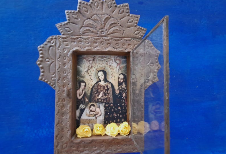 May include: A small, ornate wooden frame with a glass door. The frame is decorated with intricate metalwork and features a religious image of a woman and two children. The image is surrounded by yellow flowers.