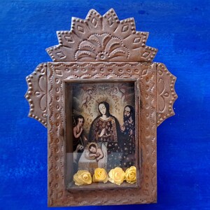 May include: A small, ornate, metal frame with a religious image inside. The frame is decorated with a scalloped top and intricate designs. The image depicts a mother and child with a man standing behind them. The image is surrounded by yellow flowers.
