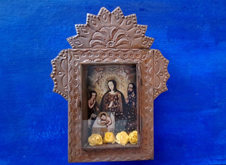 May include: A small, ornate, metal frame with a religious image inside. The frame is decorated with a scalloped top and intricate designs. The image depicts a mother and child with a man standing behind them. The image is surrounded by yellow flowers.