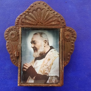 Puede incluir: Un marco de metal oxidado con una ventana de vidrio que contiene una foto antigua de un hombre con una túnica blanca. El marco tiene un diseño decorativo con un borde festoneado.