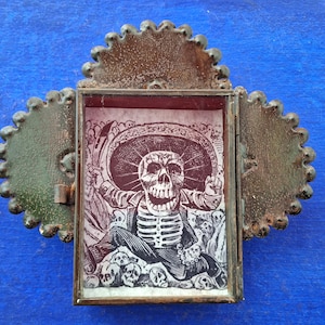 May include: A small, ornate metal frame with a hinged door, containing a printed image of a skeleton wearing a sombrero and surrounded by skulls. The frame is painted in shades of brown and green, with a red background for the image.