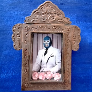 May include: A framed image of a person wearing a blue mask and a white suit. The frame is made of metal and has a decorative design. There are pink flowers in the bottom of the frame.