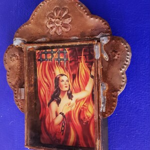 May include: A framed image of a woman in a fiery setting, with a chain around her wrist. The frame is made of rusty metal with a decorative floral design at the top.