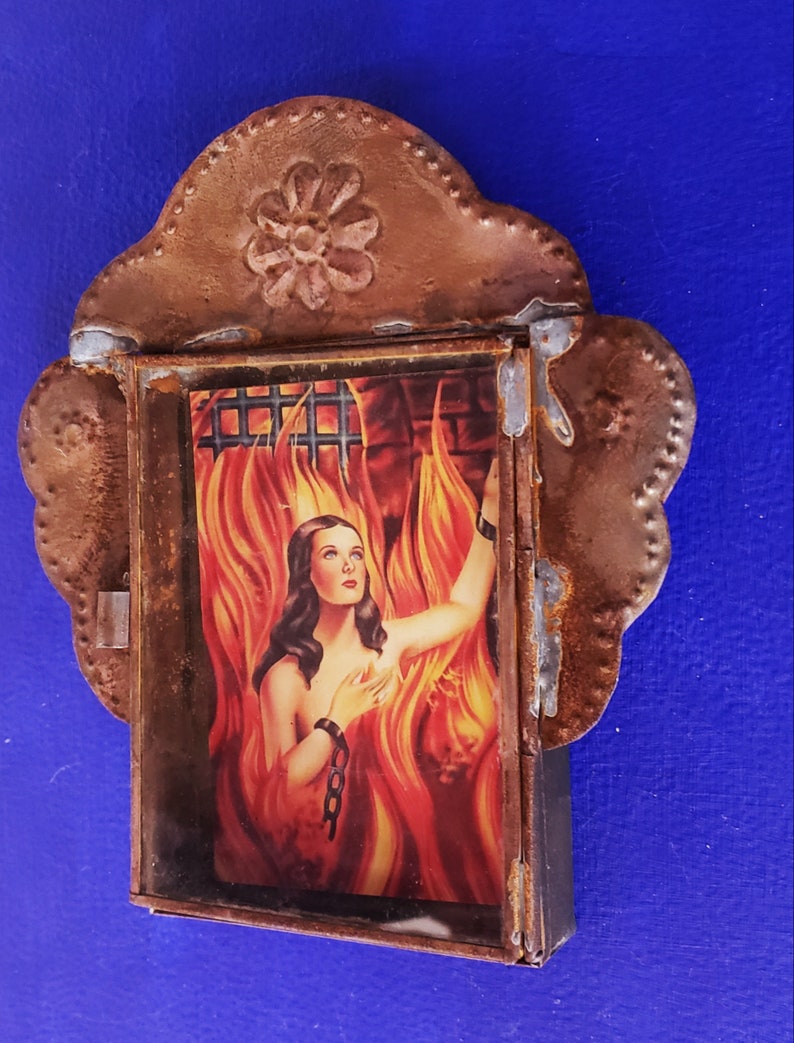 May include: A framed image of a woman in a fiery setting, with a chain around her wrist. The frame is made of rusty metal with a decorative floral design at the top.