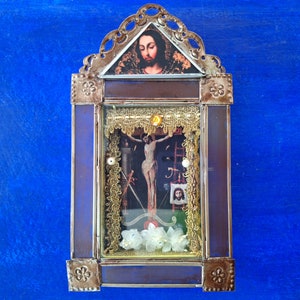 May include: A small, ornate, metal framed picture with a glass front. The picture contains a religious image of Jesus on the cross with white flowers and gold trim.