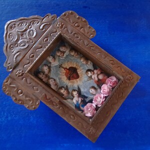May include: A small, ornate, wooden frame with a religious image inside. The image depicts a heart with a crown of thorns and angels surrounding it. The frame is decorated with pink flowers.