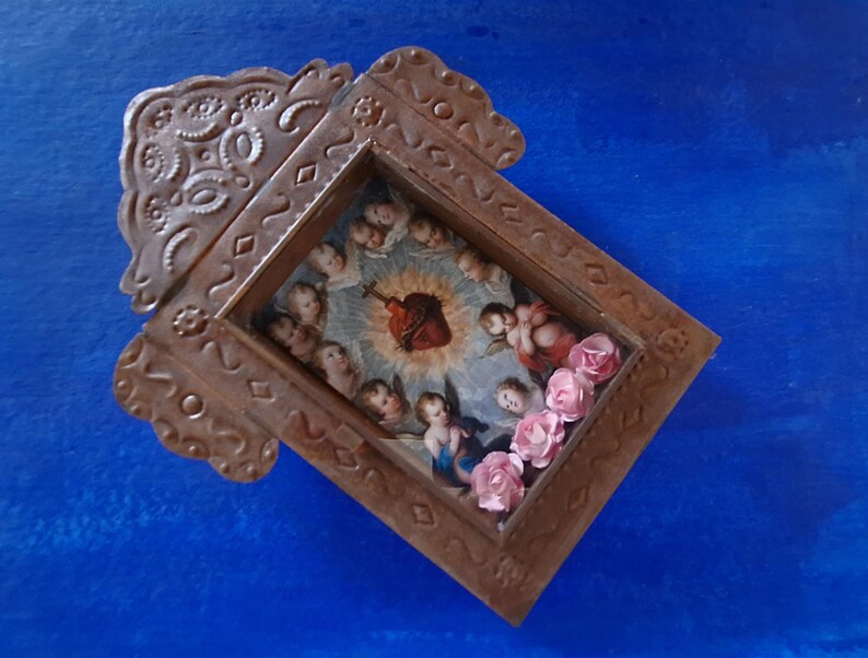 May include: A small, ornate, wooden frame with a religious image inside. The image depicts a heart with a crown of thorns and angels surrounding it. The frame is decorated with pink flowers.