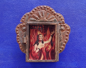May include: A small, rusty metal frame with a decorative design surrounds a picture of a woman in a red dress standing in flames. The woman is looking up and her hands are raised.