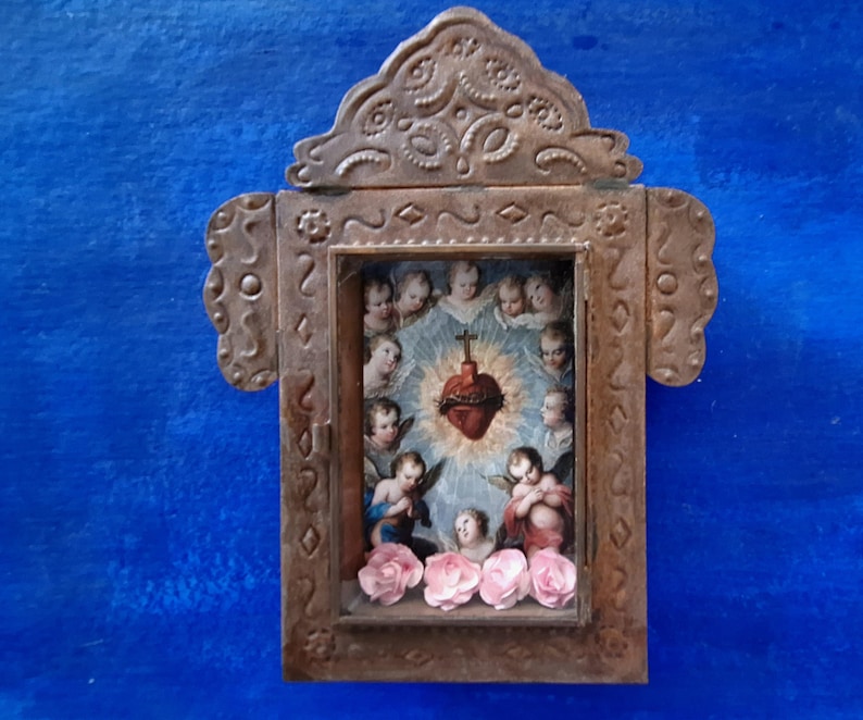 May include: A small, ornate wooden frame with a glass front, depicting a religious image of a heart with thorns and surrounded by angels. The frame is decorated with metal accents and has a blue background.