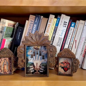 May include: A wooden bookshelf displays a collection of books and three framed religious images. The frames have ornate, textured borders. The books are of various sizes and colours, with titles visible.