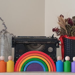 May include: A black vintage typewriter sits between a clear textured glass vase with lavender and a black vase with dried flowers. In front are six wooden peg dolls in rainbow colours and a nested wooden rainbow arch toy.