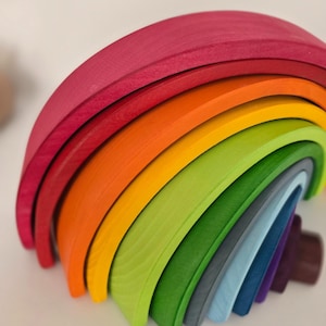 May include: A multi-colored wooden stacking toy shaped like a rainbow, consisting of nested arched pieces in shades of magenta, orange, yellow, lime green, forest green, grey, light blue, dark blue, and deep purple with a visible wood grain.