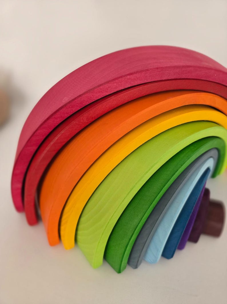 May include: A multi-colored wooden stacking toy shaped like a rainbow, consisting of nested arched pieces in shades of magenta, orange, yellow, lime green, forest green, grey, light blue, dark blue, and deep purple with a visible wood grain.